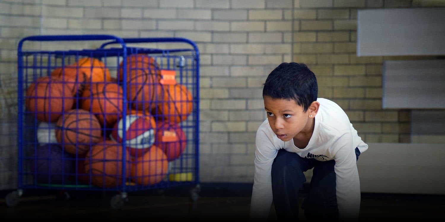 Student in a ready stance in the school gym, with a cart of basketballs in the background.
