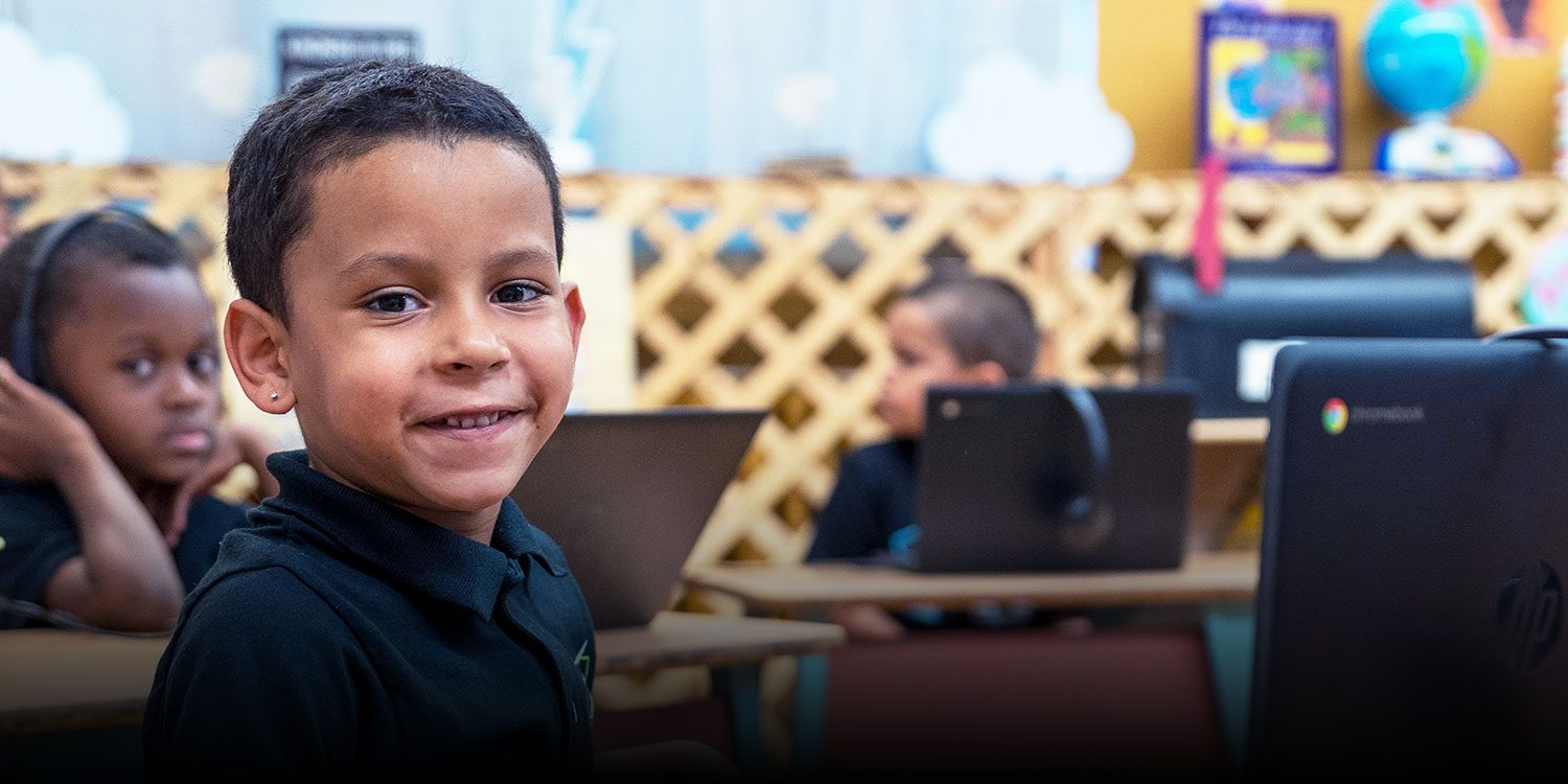 Smiling student in a classroom with classmates working on laptops.